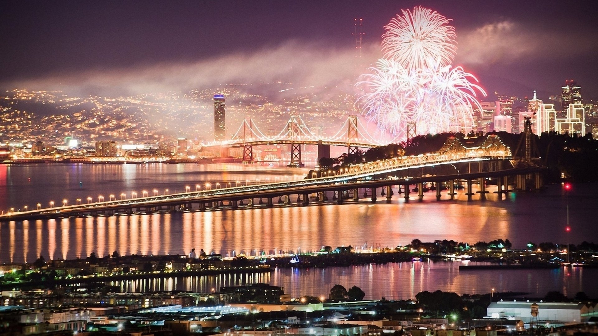 Photography of fireworks over a lit city skyline and suspension bridge at night, reflections rippling across the harbor — HD PC desktop wallpaper and background.