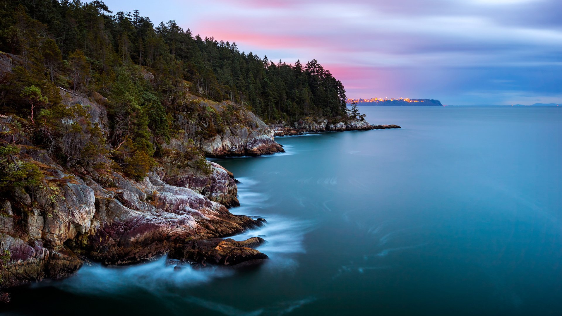 HD PC desktop wallpaper showing a serene coastline with rocky shores, dense forest, and calm blue waters under a softly colored sky at dusk.