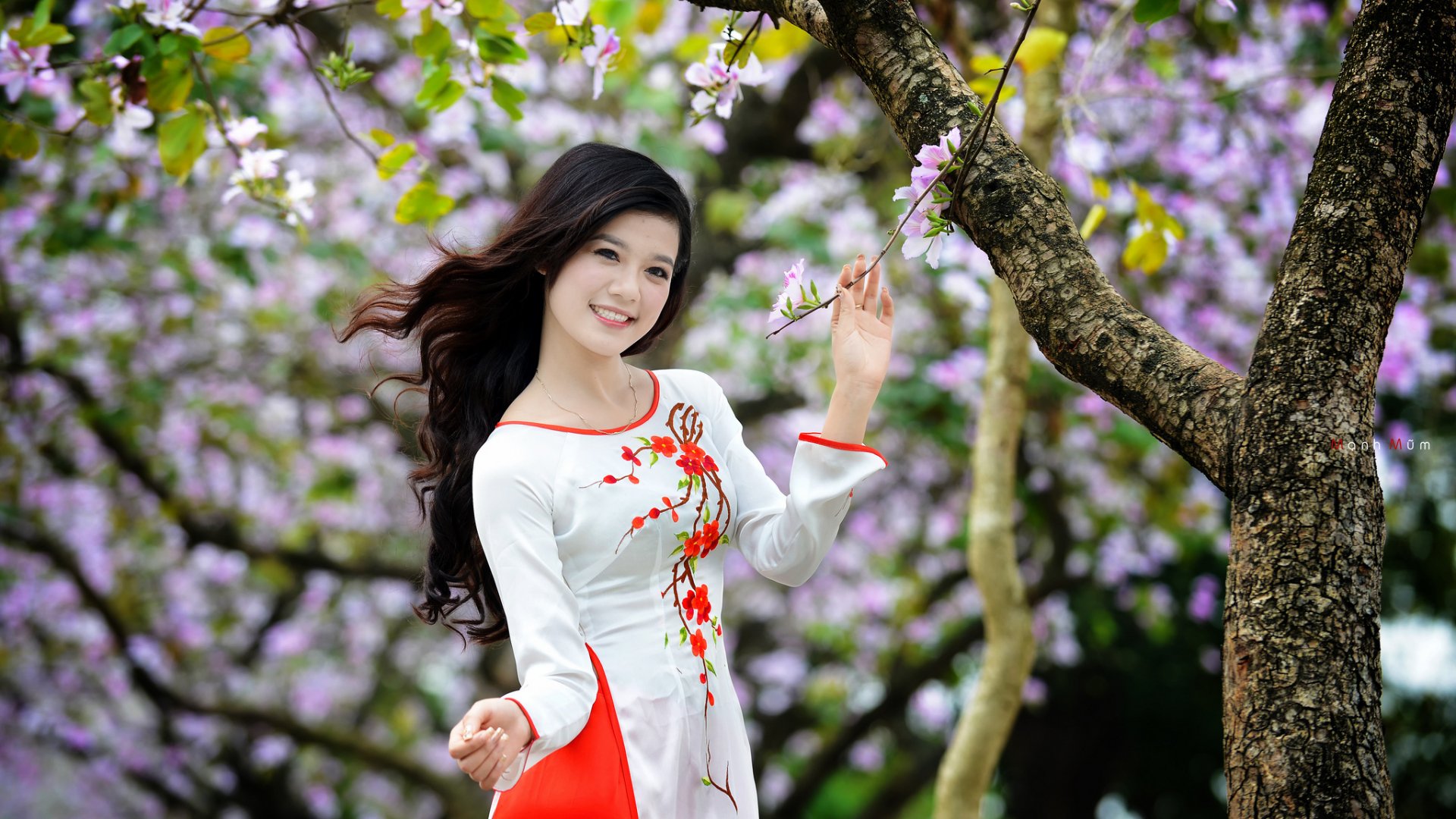 An Asian woman in a floral dress smiles while standing near a blossoming tree. The background features vibrant purple flowers, creating a serene and picturesque HD desktop wallpaper and background.