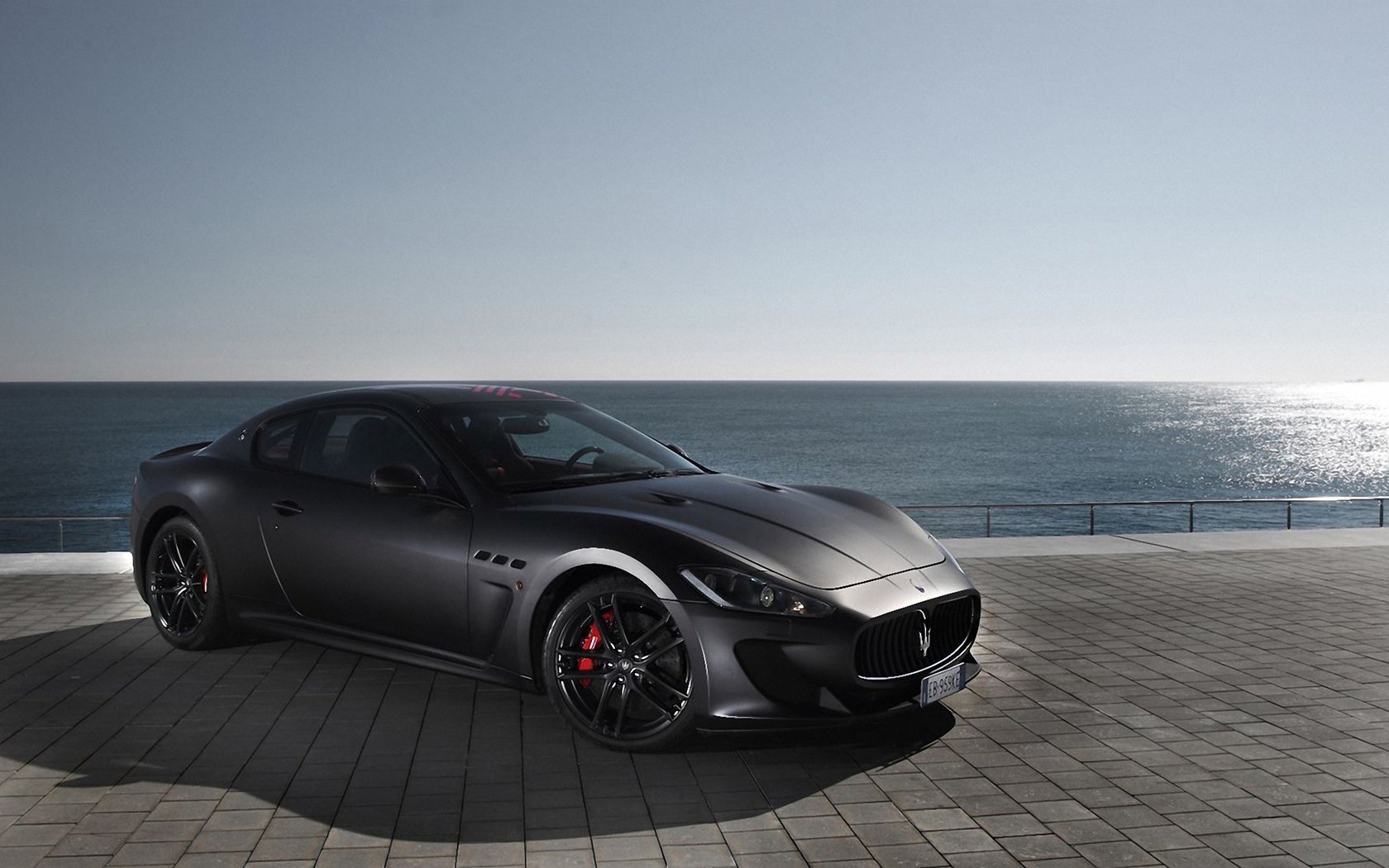 HD PC desktop wallpaper featuring a sleek black Maserati vehicle parked on a coastal promenade under a clear sky with the ocean in the background.