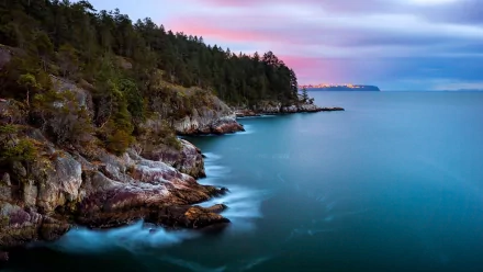 HD PC desktop wallpaper showing a serene coastline with rocky shores, dense forest, and calm blue waters under a softly colored sky at dusk.