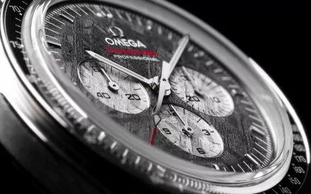 Close-up of a man-made Omega chronograph watch face with textured dial and stainless-steel case, red-tipped hand — 2K Quad HD PC desktop wallpaper/background.
