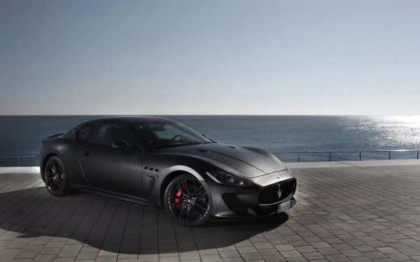 HD PC desktop wallpaper featuring a sleek black Maserati vehicle parked on a coastal promenade under a clear sky with the ocean in the background.