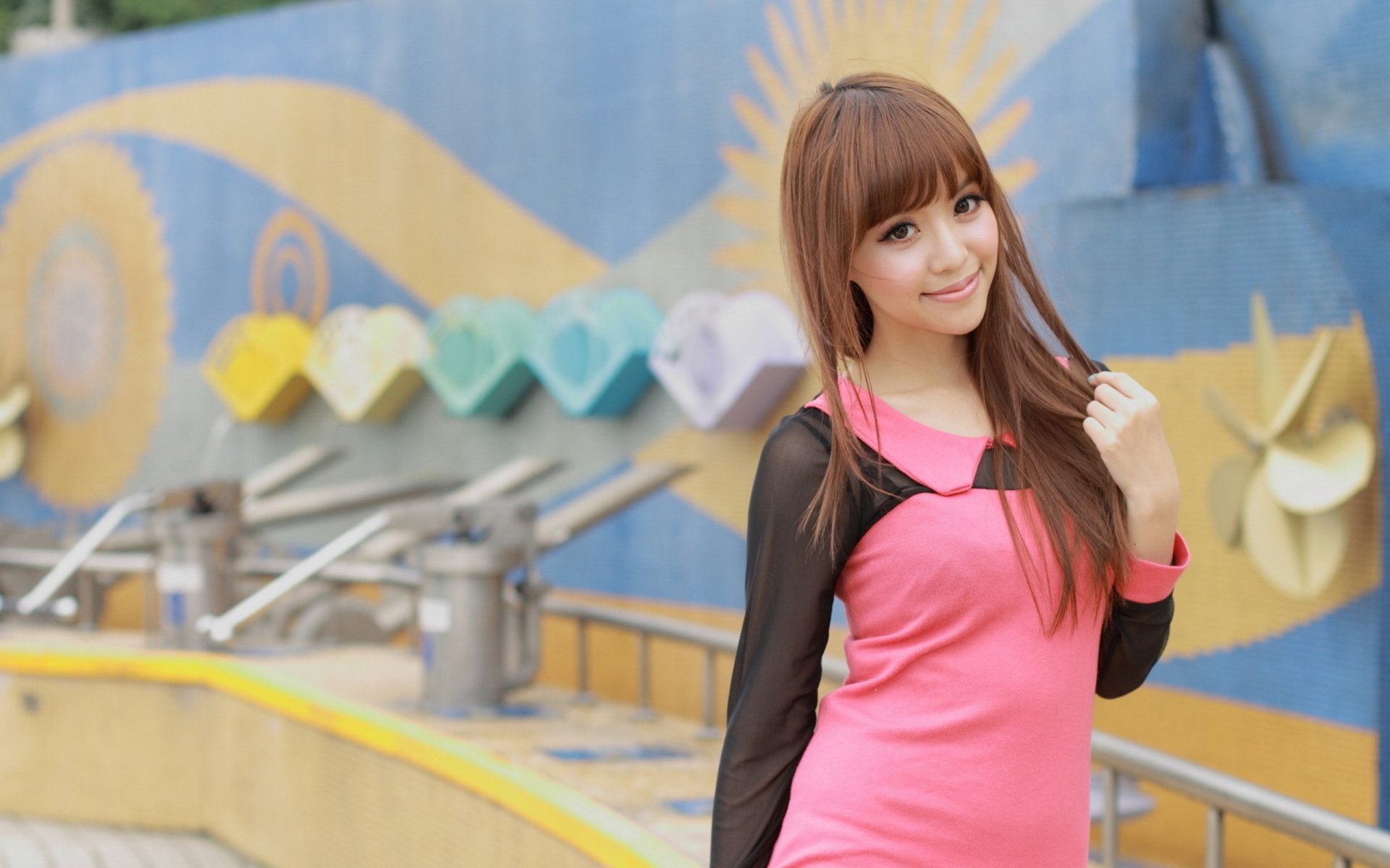 A woman of Asian descent in a pink and black outfit stands in front of a colorful outdoor backdrop featuring heart designs and wavy patterns. This is an HD desktop wallpaper and background image.