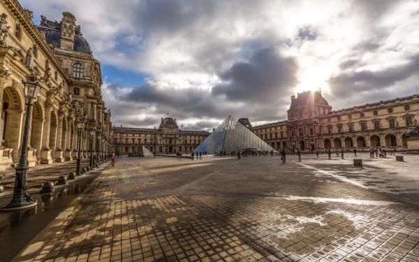 A stunning HD wallpaper showcasing the iconic Louvre Pyramid and its surrounding architecture, illuminated by dramatic clouds and sunlight in a tranquil courtyard.