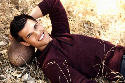 A smiling Taylor Lautner relaxes on the ground, leaning against a football, surrounded by golden grass. This striking image serves as a captivating HD desktop wallpaper.