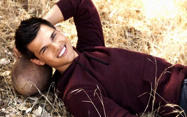 A smiling Taylor Lautner relaxes on the ground, leaning against a football, surrounded by golden grass. This striking image serves as a captivating HD desktop wallpaper.