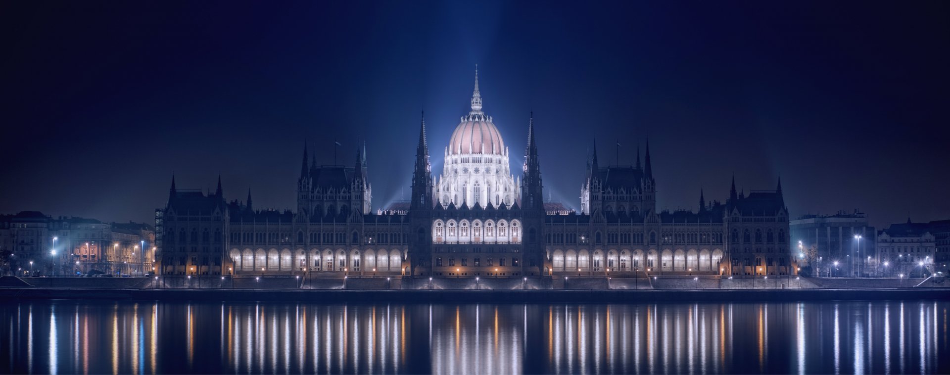 HD desktop wallpaper of the Hungarian Parliament Building illuminated at night, reflecting on the calm river with a deep blue sky background.