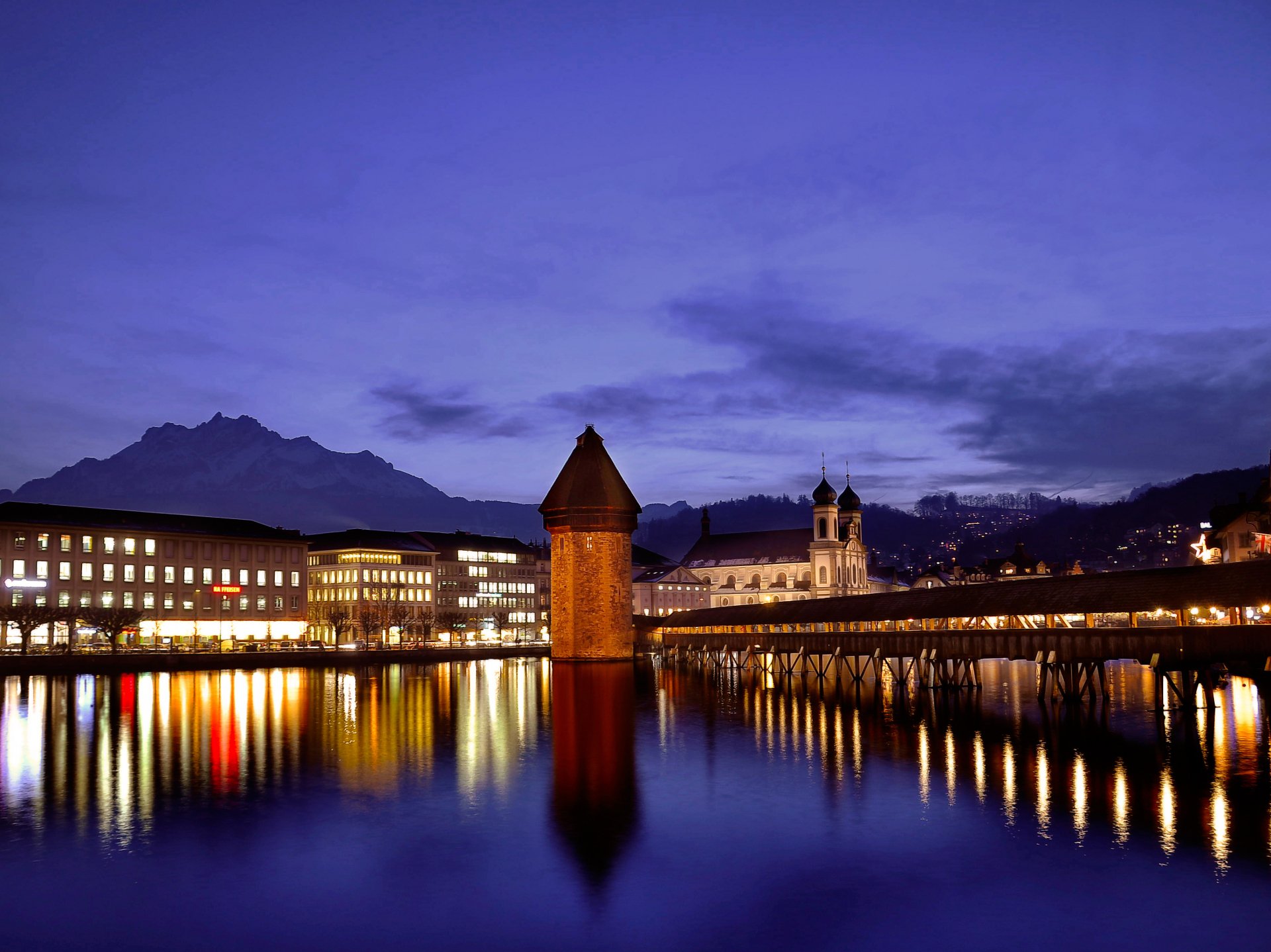HD desktop wallpaper of the man-made Chapel Bridge and Water Tower illuminated at dusk in Lucerne, reflecting beautifully on the calm lake waters.