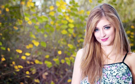 A woman with long blonde hair stands outdoors, surrounded by lush greenery and yellow leaves. The HD desktop wallpaper captures a serene mood.