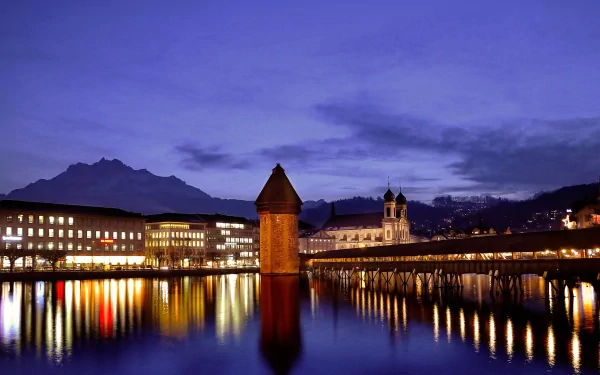 HD desktop wallpaper of the man-made Chapel Bridge and Water Tower illuminated at dusk in Lucerne, reflecting beautifully on the calm lake waters.