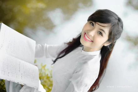 HD desktop wallpaper featuring an Asian woman in a white blouse, smiling while holding an open book, with a blurred natural background.
