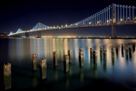 Night view of the Bay Bridge spanning San Francisco's ocean shoreline in California, with illuminated cables reflecting on calm water and weathered wooden posts in the foreground.