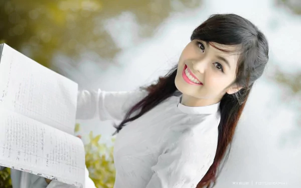 HD desktop wallpaper featuring an Asian woman in a white blouse, smiling while holding an open book, with a blurred natural background.
