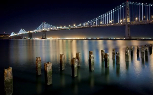 Night view of the Bay Bridge spanning San Francisco's ocean shoreline in California, with illuminated cables reflecting on calm water and weathered wooden posts in the foreground.