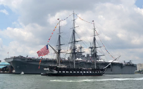HD PC desktop wallpaper background: a modern military ship escorts a tall, flag-flying sailing vessel on calm water beneath a cloudy sky.