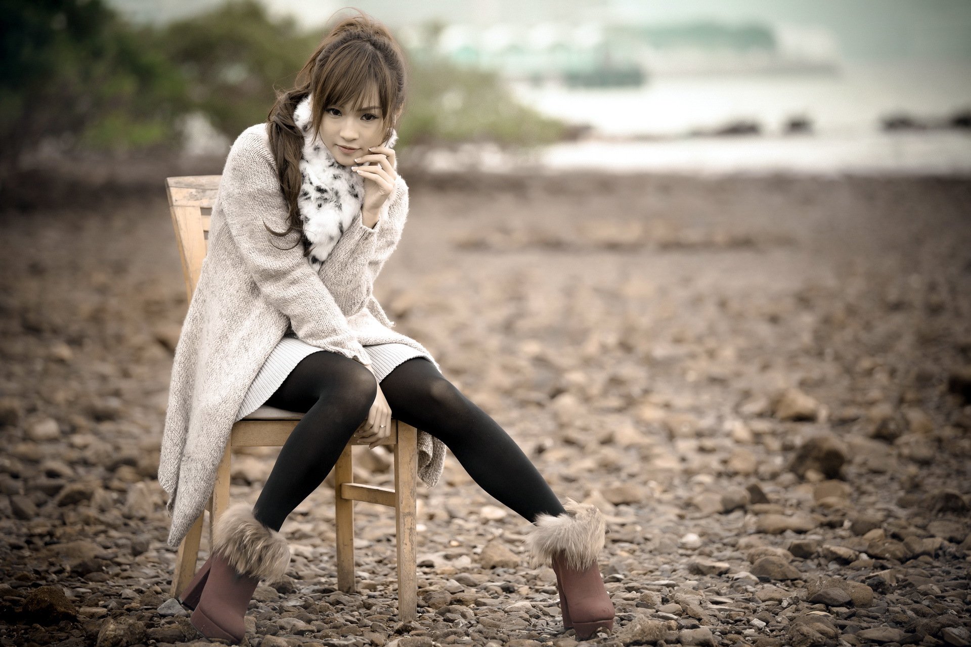 A woman sits on a chair in an outdoor setting, wearing a coat, scarf, and fur-lined boots. The background features a rocky terrain. Asian woman, HD desktop wallpaper and background.