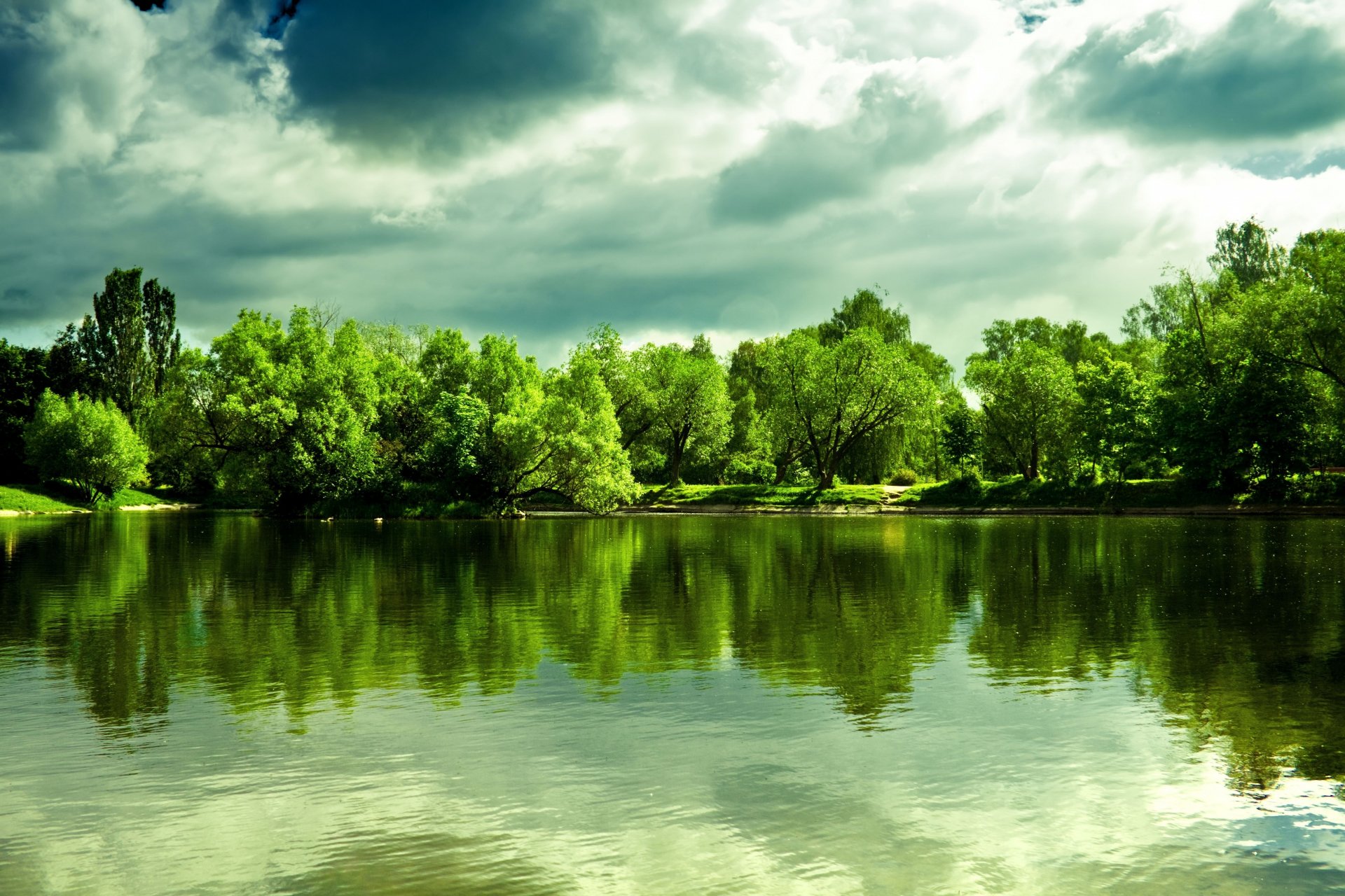 Lush greenery and dramatic clouds reflect beautifully on a serene lake in this vibrant nature scene, showcasing stunning 4K Ultra HD detail for a captivating desktop wallpaper.