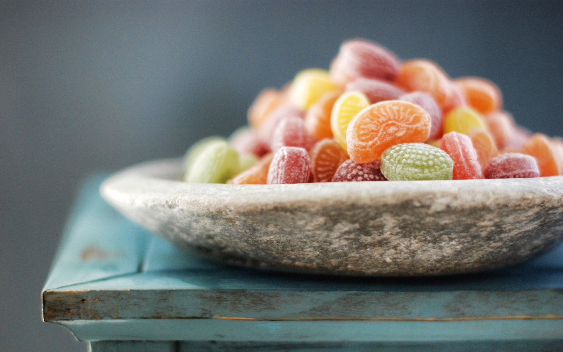 A vibrant assortment of colorful candies is displayed in a rustic bowl on a teal wooden table, creating a cheerful and inviting food-themed desktop wallpaper.