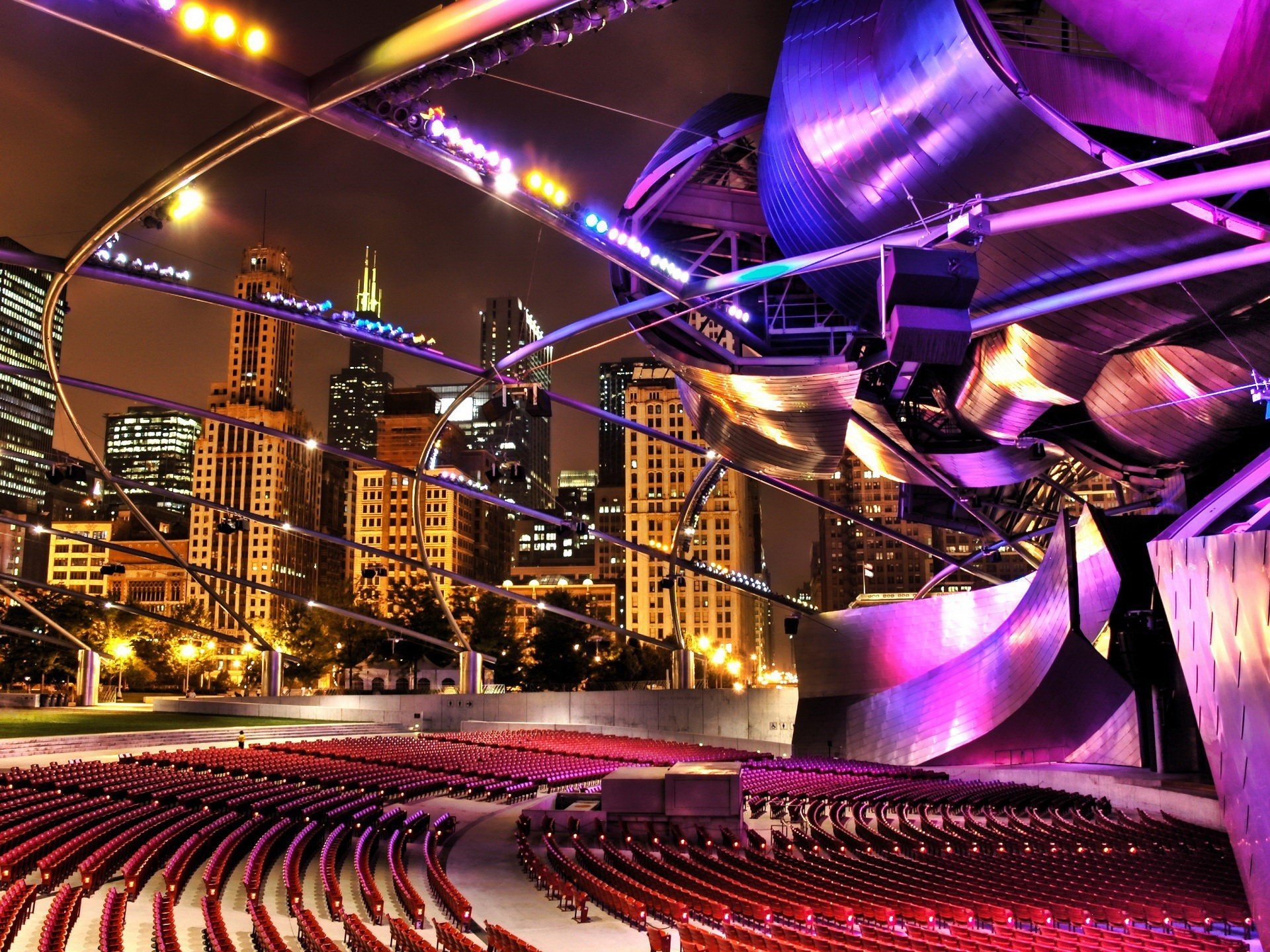 HD desktop wallpaper of Chicago's illuminated outdoor theater with sweeping metallic stage, empty seats and a glowing city skyline at night — man-made lights and architecture.