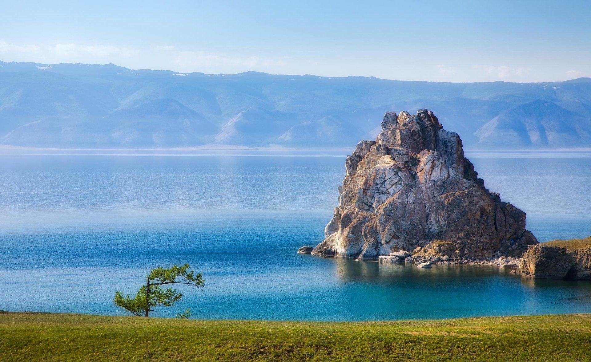 Scenic HD desktop wallpaper featuring a serene mountain landscape with a rocky island surrounded by a calm lake and a grassy foreground under a clear sky.