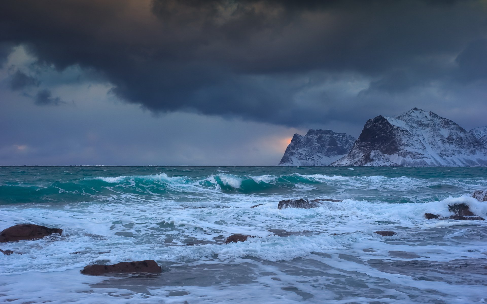 HD PC desktop wallpaper of turbulent ocean waves crashing on rocky shore under a moody cloudy sky with distant mountainous coastline, showcasing nature's raw beauty.