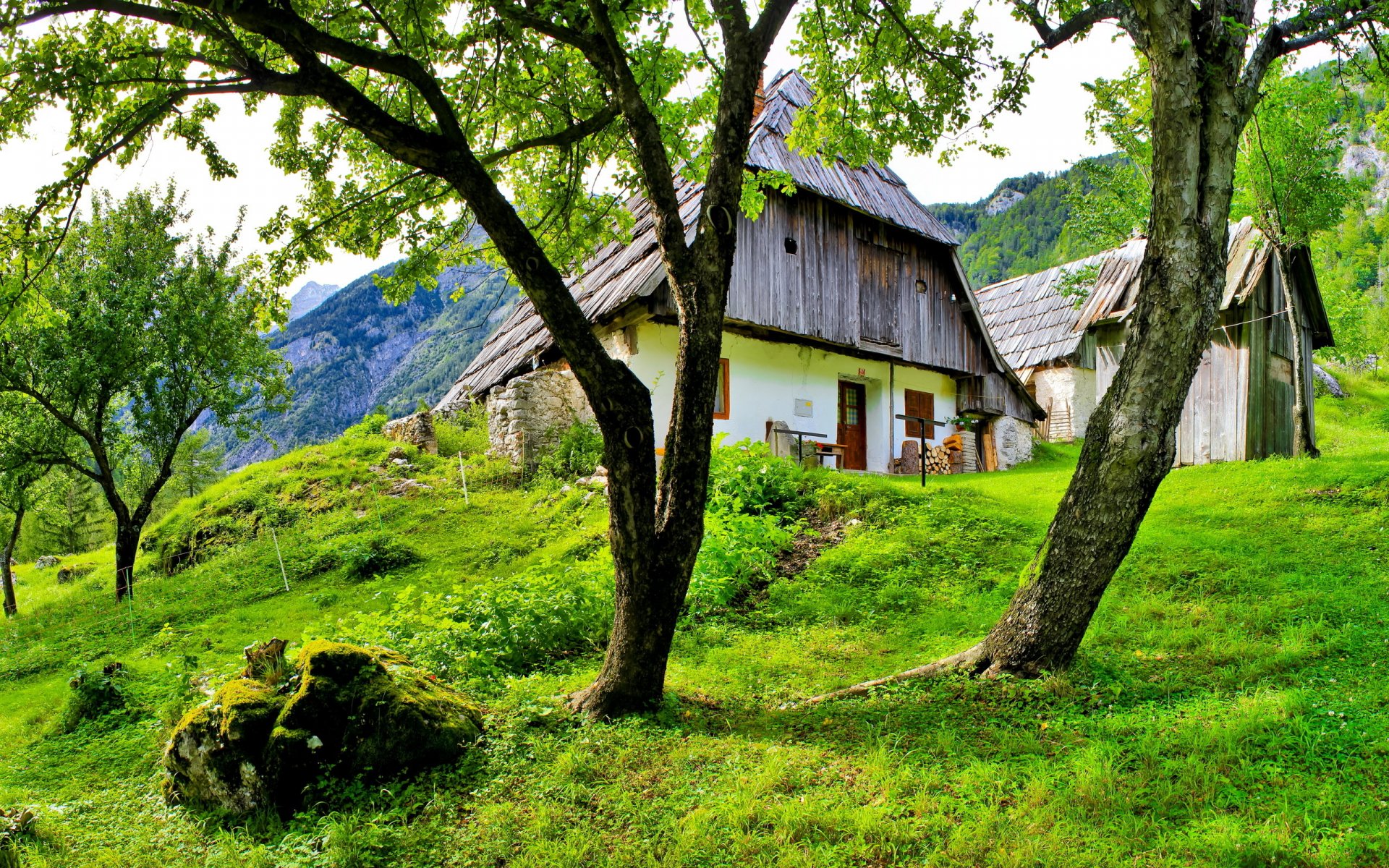 HD desktop wallpaper featuring a man-made rustic cabin nestled among trees on a lush, green hillside with mountains in the background.