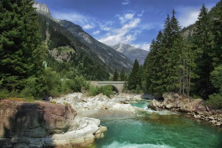 HD desktop wallpaper featuring a man-made stone bridge spanning a clear river, surrounded by lush pine trees and towering mountain peaks under a bright blue sky.