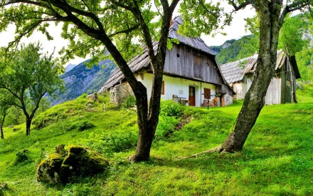 HD desktop wallpaper featuring a man-made rustic cabin nestled among trees on a lush, green hillside with mountains in the background.