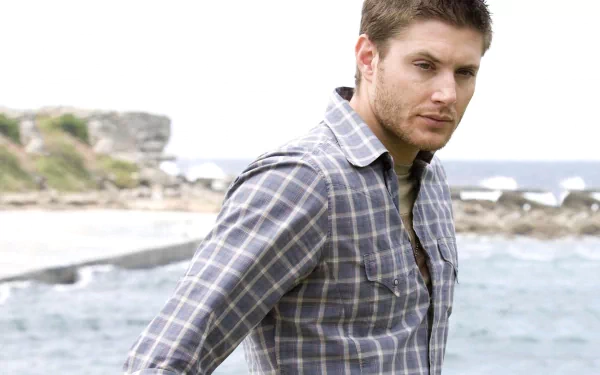 HD PC desktop wallpaper featuring celebrity Jensen Ackles standing by a rocky coastline, wearing a plaid shirt and looking thoughtfully into the distance.