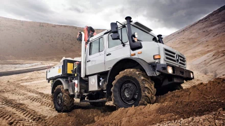 HD PC desktop wallpaper showing a Mercedes-Benz Unimog vehicle navigating muddy off-road terrain beneath a dramatic cloudy sky