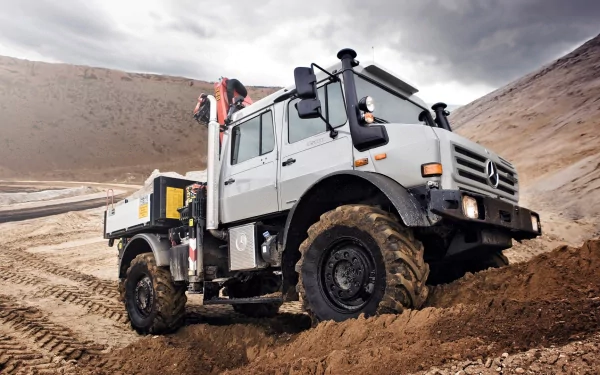 HD PC desktop wallpaper showing a Mercedes-Benz Unimog vehicle navigating muddy off-road terrain beneath a dramatic cloudy sky