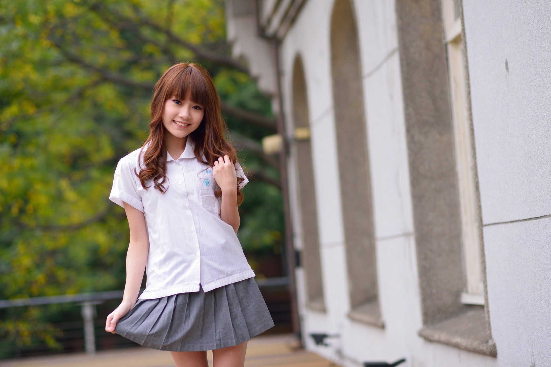 HD desktop wallpaper showing an Asian woman with long brown hair, dressed in a white shirt and gray skirt, standing outdoors near a building with archways and lush green trees in the background.