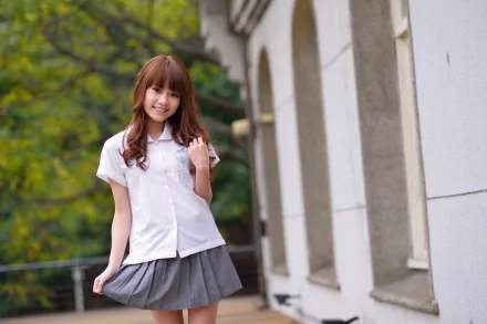 HD desktop wallpaper showing an Asian woman with long brown hair, dressed in a white shirt and gray skirt, standing outdoors near a building with archways and lush green trees in the background.