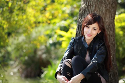 Asian woman sitting outdoors, leaning against a tree, smiling. She is wearing a black leather jacket. Lush green foliage in the background. HD desktop wallpaper and background.