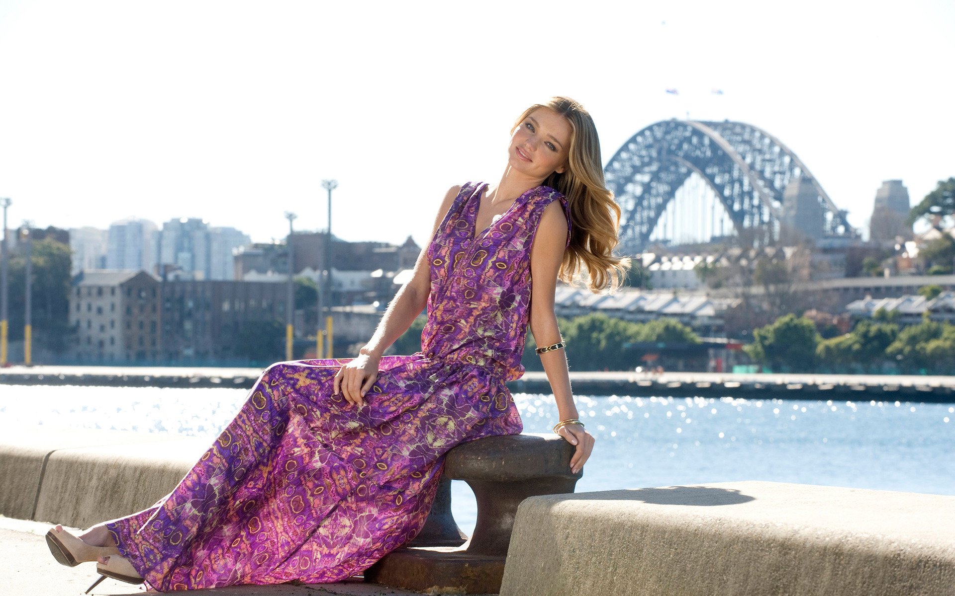 Miranda Kerr sits elegantly in a vibrant purple dress by the waterfront, with the Sydney Harbour Bridge in the background, creating a stunning HD desktop wallpaper.