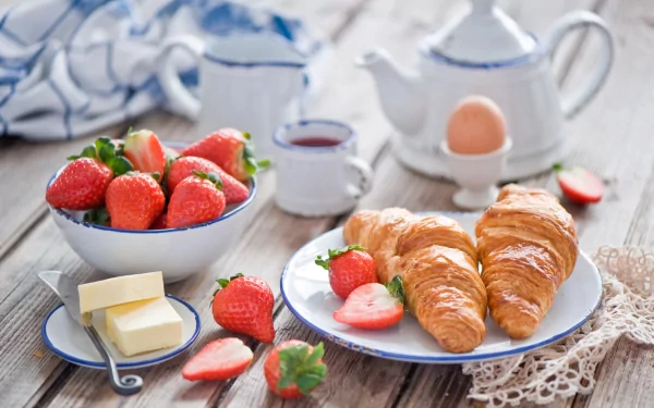A vibrant still life showcasing a delicious breakfast, featuring fresh strawberries, butter, a perfectly cooked egg, croissants, and a teapot, set against a rustic wooden table.