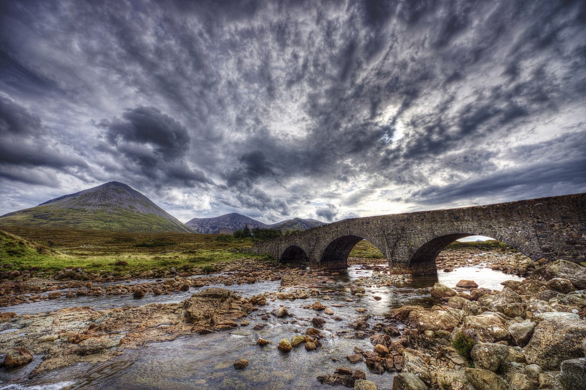 HD PC desktop wallpaper background showing a man-made stone arch bridge crossing a rocky stream beneath dramatic stormy clouds and distant rolling hills.