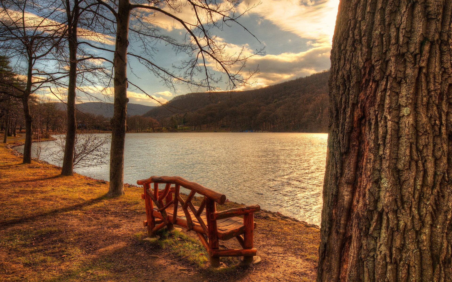 HD desktop wallpaper depicting a man-made wooden bench beside a calm lake, framed by trees and a glowing sunset sky.