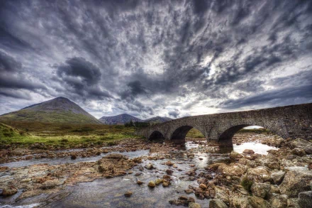 HD PC desktop wallpaper background showing a man-made stone arch bridge crossing a rocky stream beneath dramatic stormy clouds and distant rolling hills.