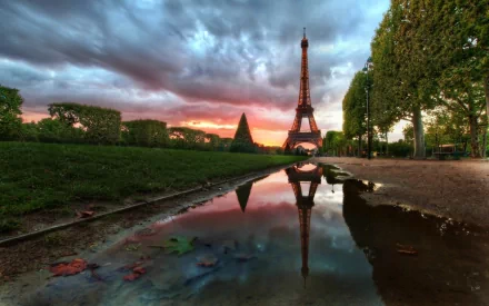 HD wallpaper showing the Eiffel Tower at sunset, with dramatic clouds and a lush green landscape. The Eiffel Tower and the colorful sky are beautifully reflected in a puddle on the path.