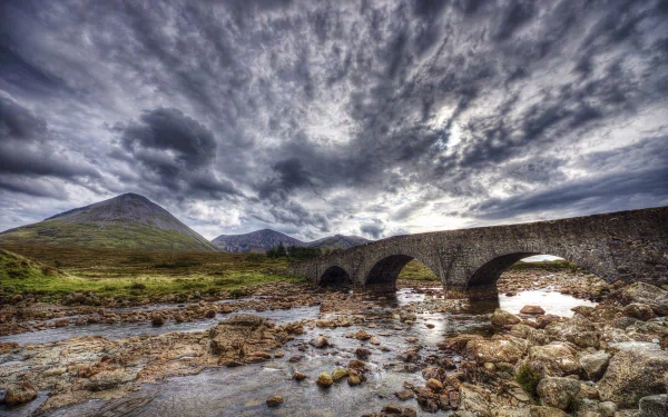 HD PC desktop wallpaper background showing a man-made stone arch bridge crossing a rocky stream beneath dramatic stormy clouds and distant rolling hills.