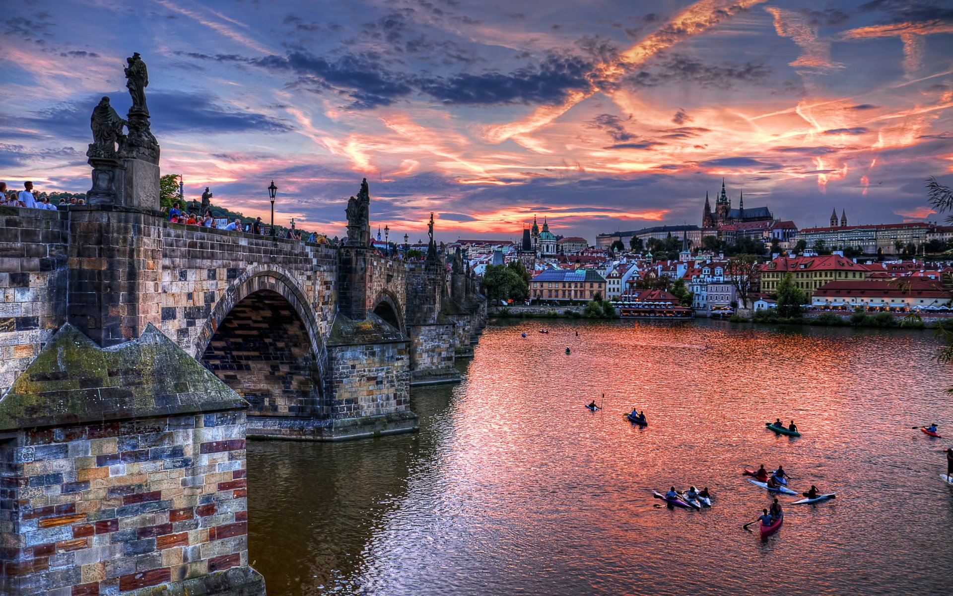 HD desktop wallpaper showcasing the historic Charles Bridge and Prague skyline at sunset with boats on the Vltava River.