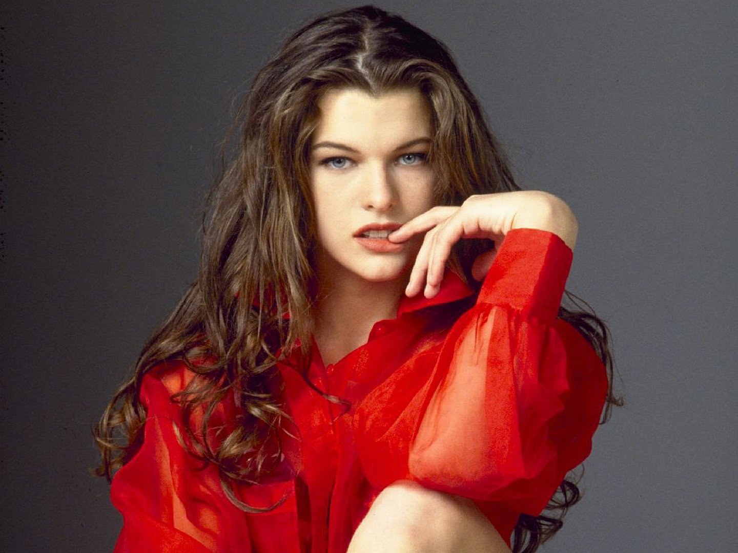 HD PC desktop wallpaper of celebrity Milla Jovovich wearing a red sheer blouse, posing thoughtfully against a plain dark background.