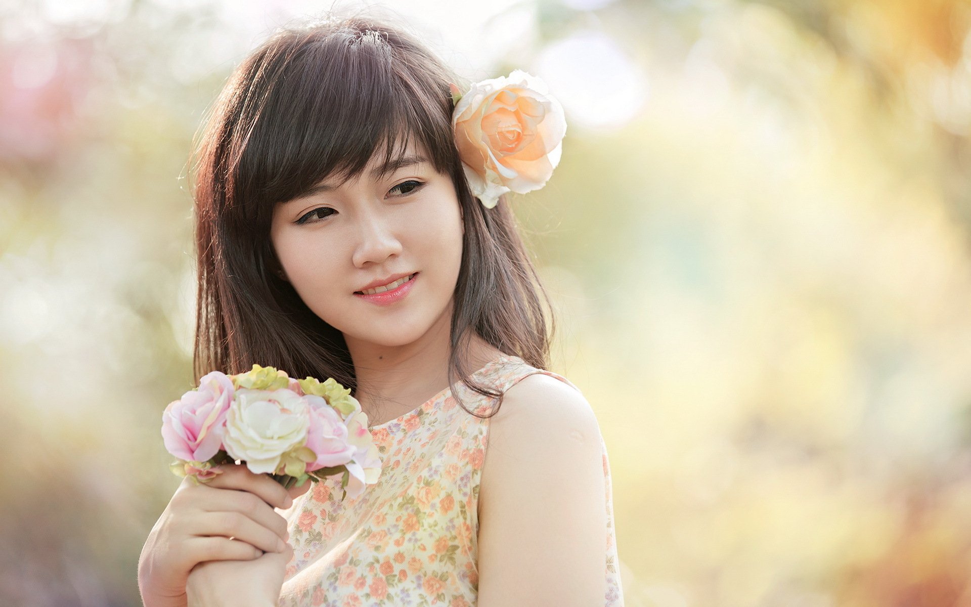 HD desktop wallpaper featuring a smiling Asian woman with long dark hair, holding a bouquet of flowers, and a peach rose in her hair against a soft, blurry floral background.
