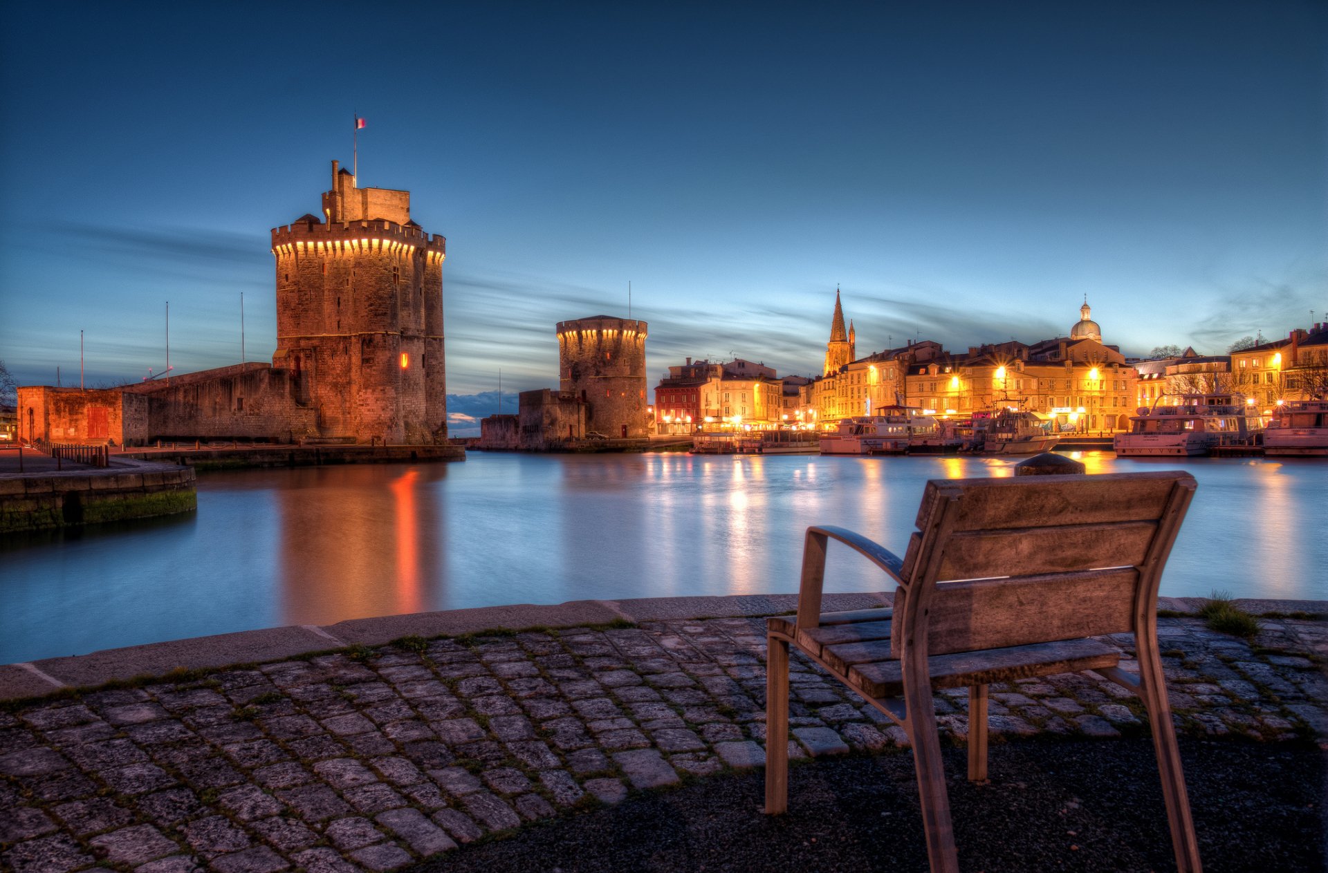 HD desktop wallpaper showcasing a man-made town at dusk with historic towers, waterfront buildings, and a wooden bench in the foreground.
