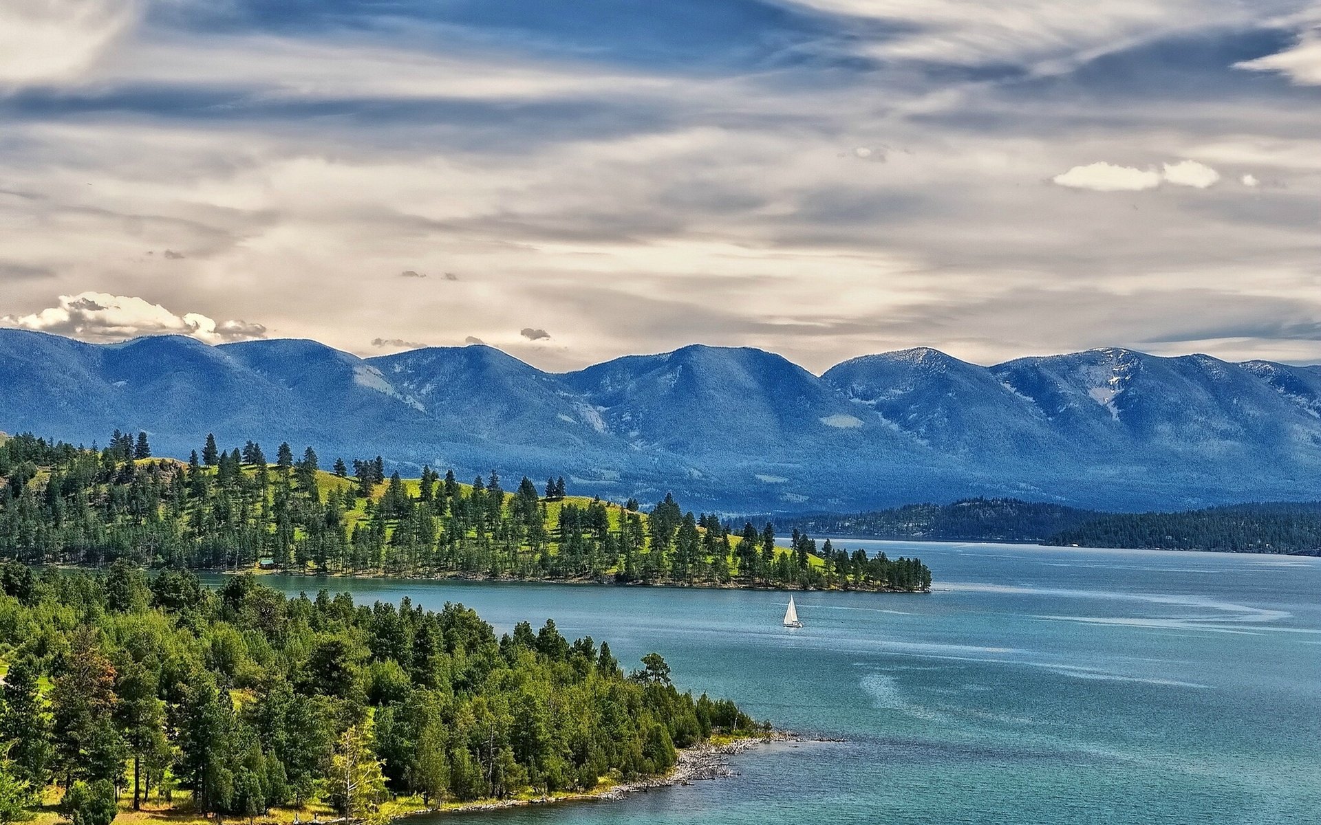 HD PC desktop wallpaper showcasing a serene nature landscape with a forested shoreline, expansive lake, sailboat, and mountains under a cloudy sky.