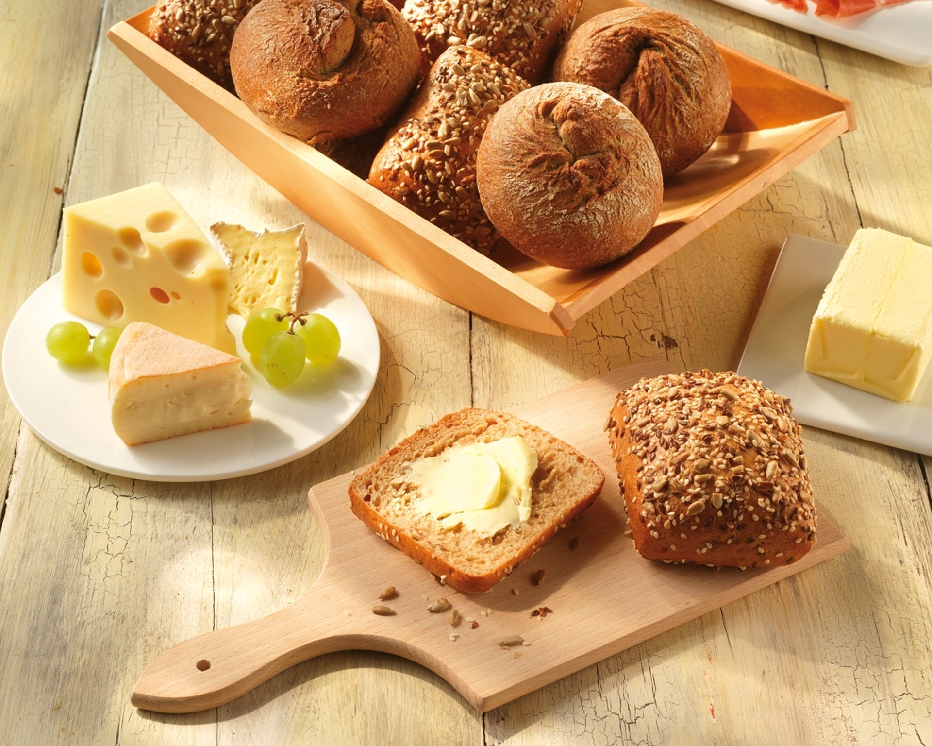 HD desktop wallpaper showing an assortment of bread rolls, slices of cheese, butter, and grapes arranged on a wooden table.