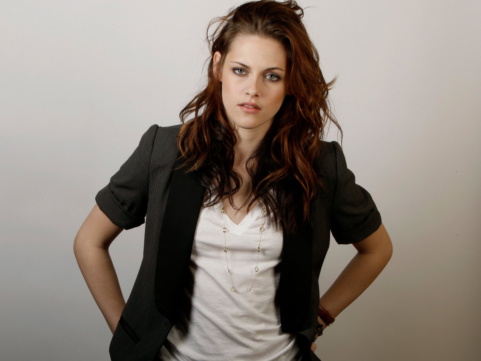 HD desktop wallpaper featuring celebrity Kristen Stewart. She stands with hands on hips, wearing a grey blazer over a white shirt, with a neutral expression against a plain background.