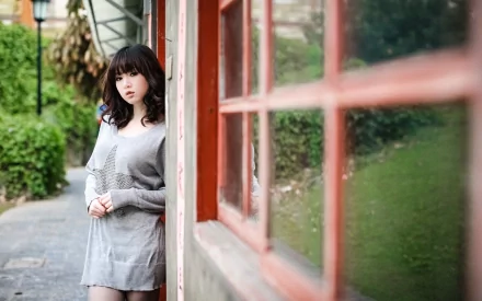 HD desktop wallpaper of an Asian woman standing outdoors by a building with red-framed windows, surrounded by greenery.